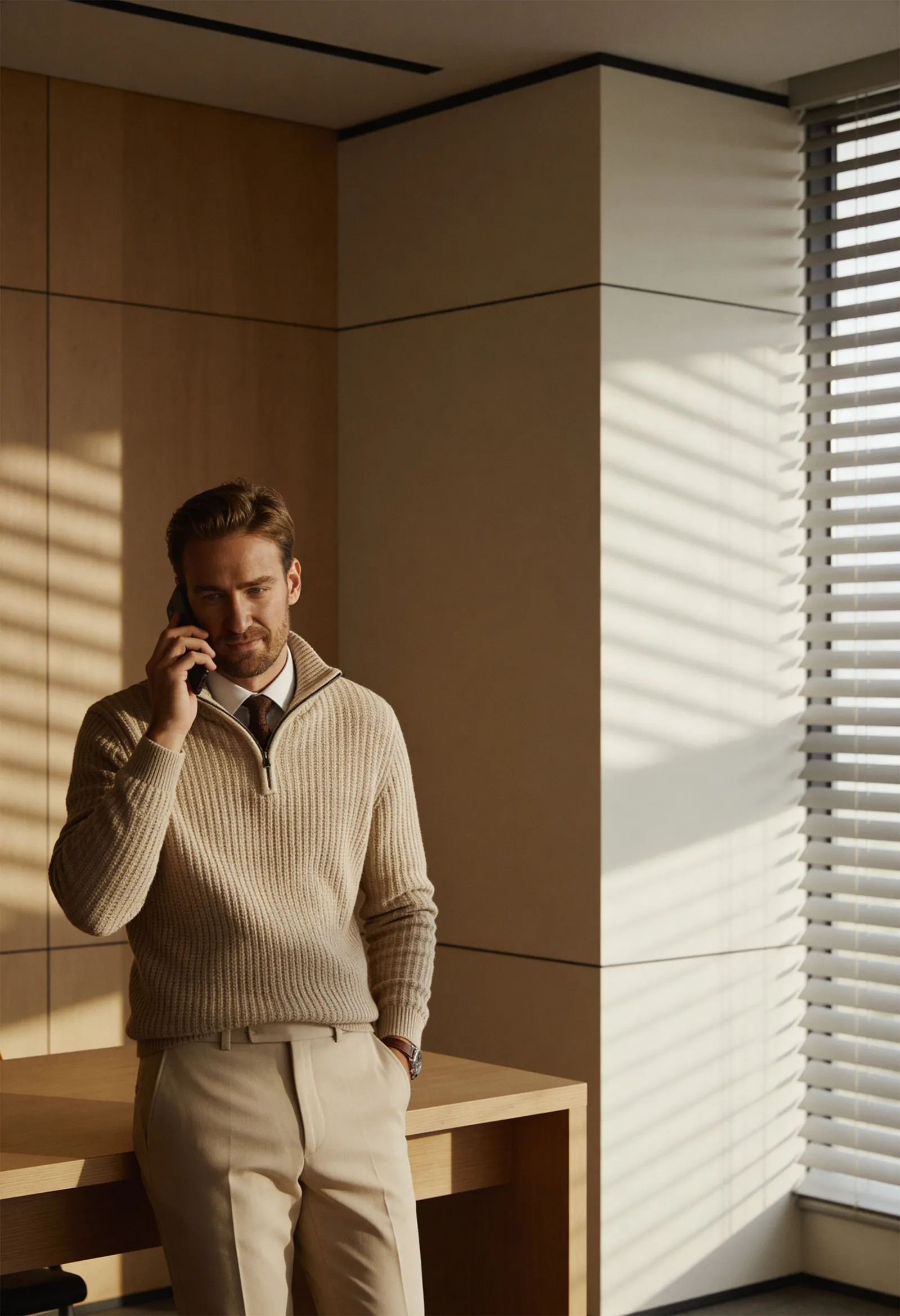 Man in beige sweater and dress pants standing by a wooden desk, talking on a phone in an office with sunlight coming through window blinds.